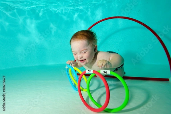 Obraz A happy kid with Down syndrome swims and collects toys underwater from the bottom of a children's pool during a sports activity. Children's disability. Concept. Portrait. Horizontal orientation.