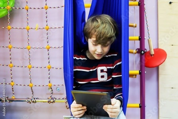 Fototapeta child boy watching cartoons on a tablet while sitting in a hammock at a children's center for the development of special children