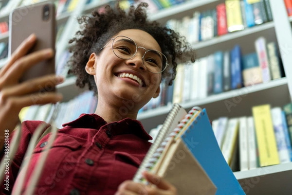 Fototapeta Young and beautiful businesswoman holding a lot of books in her hands and standing near the bookshelves in the modern office and using smartphone with wide smile