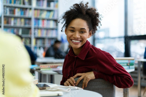 Fototapeta Waist up portrait view of the multiracial woman looking at the camera with toothy smile while spending time at the school library