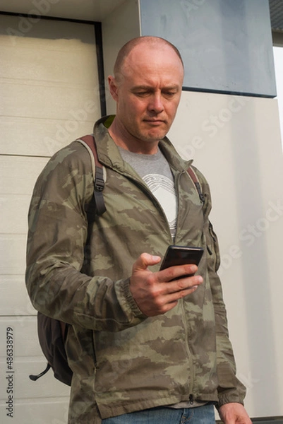 Fototapeta A sporty man in a military jacket with a mobile phone in his hands sends a message on the street