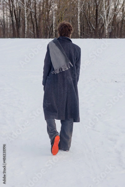 Fototapeta A young man in a long gray coat and scarf walks through the snow. Rear view. Back view.