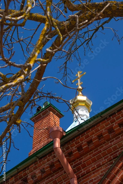 Fototapeta An Orthodox cross shining in the sun on the dome of the church. The dome is a brick building. In the foreground are blurred dry branches of a tree without leaves. Bottom view. Daylight. Spring.