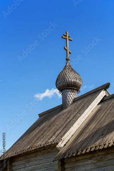 Fototapeta The wooden roof of the Orthodox chapel with a small wooden dome and a cross on it. Carved. Gray tree. Folk architecture. Clear blue sky. Daylight.