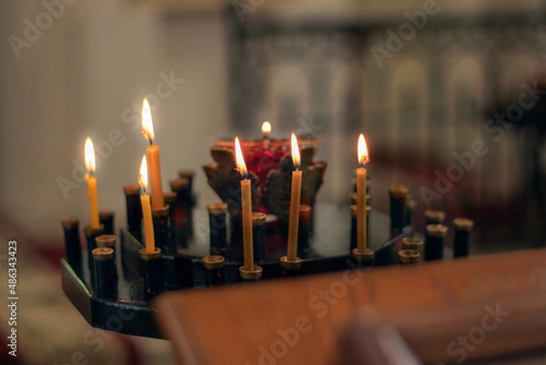 Fototapeta Burning candles in a black church candlestick. Blurred background and foreground. There is a blurred wooden pedestal in the foreground. Close-up.