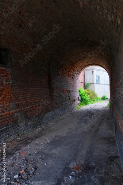 Fototapeta The tunnel of the gateway of the old red brick house. A tunnel with an arched vault. The old courtyard can be seen in the distance. Summer. Daylight.