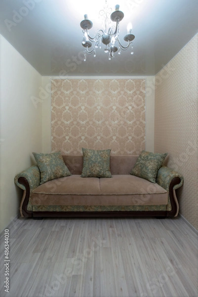 Fototapeta A green-beige classic sofa with pillows in a room with a chandelier. The sofa is surrounded on three sides by walls. Wallpaper in moldings. Stretch ceiling. Laminate flooring on the floor.
