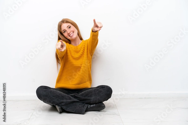 Fototapeta Young redhead woman sitting on the floor isolated on white background points finger at you while smiling