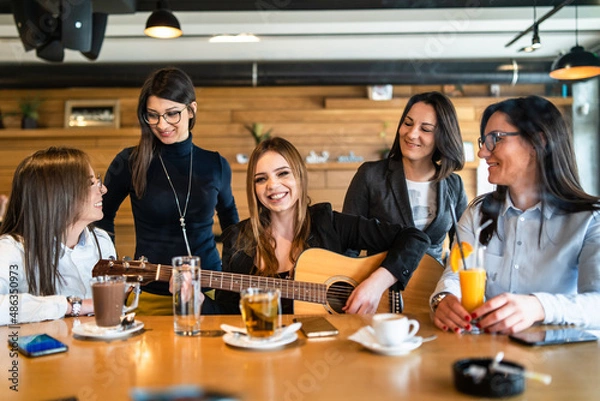 Fototapeta Young millennial women sitting by the table at cafe or restaurant singing and smiling having fun with guitar - Beautiful caucasian women having a good time while playing guitar - friendship concept
