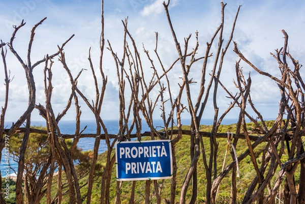 Obraz A wooden fence built of dry branches against the background of a green hill and blue sea. On the fence a blue sign with the Italian text "proprieta privata" which means "private property"
