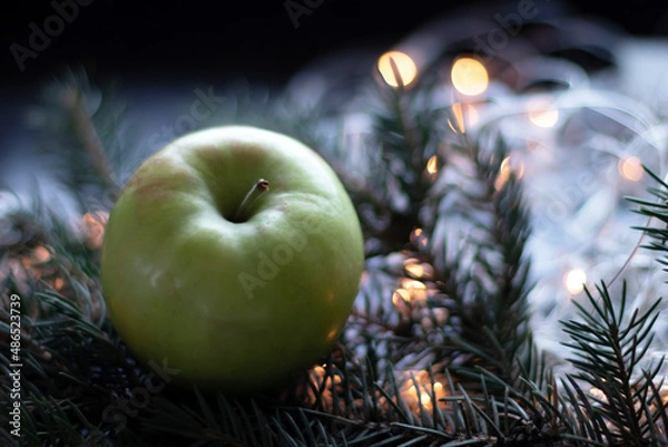 Fototapeta Green apple on Christmas tree branch. Bokeh background with garland. The concept of healthy eating during the New Year holidays
