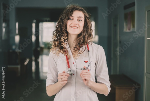 Fototapeta A woman doctor in a protective mask and a white coat stands against the backdrop of a polyclinic