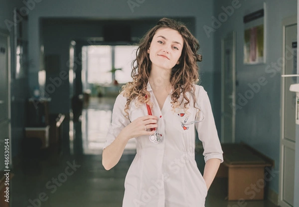 Fototapeta A woman doctor in a protective mask and a white coat stands against the backdrop of a polyclinic