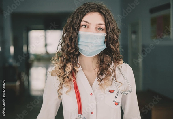Fototapeta A woman doctor in a protective mask and a white coat stands against the backdrop of a hospital