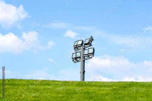 Fototapeta Electric spotlight on a metal pole during a sunny day on a green lawn. White clouds in the blue sky in the background