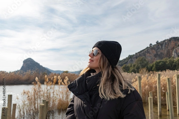 Fototapeta Happy tourist girl in front of reeds. River at background. She has sun glasses and hat. 