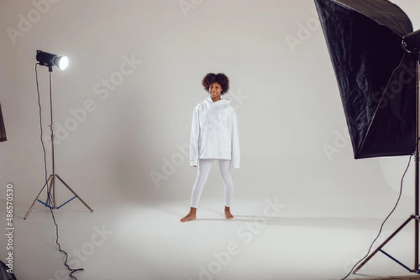 Fototapeta African American woman with puffy hair in a white hoodie and leggings stands on a white background in a photo studio.