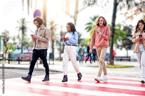 Fototapeta Group of smiling friends walking on the crosswalk and looking at the smartphone - Millennials people have fun together down the city street - Young university students stroll towards the campus