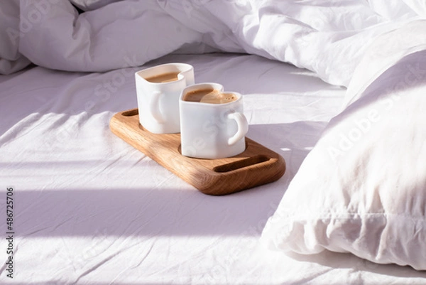 Obraz Coffee in mugs in the shape of a heart on a tray lie on a white bed. In the morning, the sun's rays illuminate the cups and a crumpled white sheet.