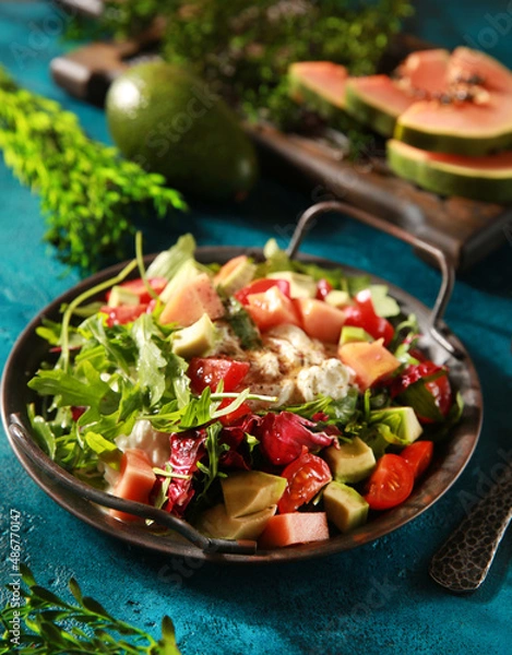 Fototapeta Tropical salad on a bright turquoise background. Fresh salad in a gray plate of arugula, papaya, avocado, cherry tomatoes, strachatella and pine nuts. Exotic dish. Background image, copy space