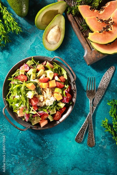 Fototapeta Tropical salad on a bright turquoise background. Fresh salad in a gray plate of arugula, papaya, avocado, cherry tomatoes, strachatella and pine nuts. Exotic dish. Background image, copy space