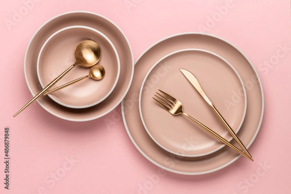Obraz Dishes and utensils for serving and eating meals. Beige round rimmed plates and golden cutlery on a pink background, top view. Modern craft ceramic crockery, trendy tableware from above.