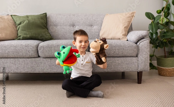 Fototapeta a Caucasian little boy is sitting on the carpet near the sofa and playing a puppet theater with a monkey and a frog
