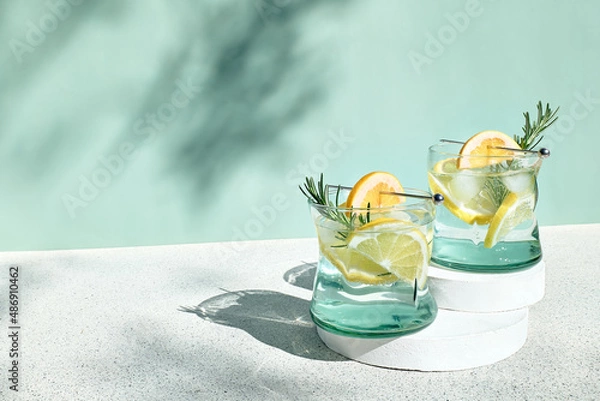 Fototapeta Summer refreshing lemonade drink or alcoholic cocktail with ice, rosemary and lemon slices on pastel light green surface. Fresh healthy cold lemon beverage. Water with lemon.