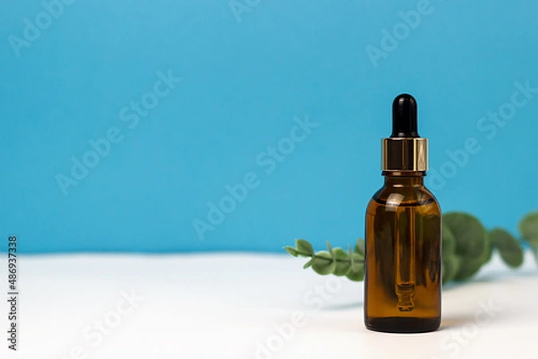 Fototapeta brown glass frosted cosmetic jar for cream and with a dropper on a blue and white background. Eucalyptus branches. Place for notes and text. Personal care and cosmetology.