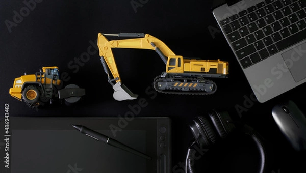 Fototapeta Stylus and graphics tablet, pc mouse, laptop and headphones on black next to models excavator and asphalt roller. Сoncept of designing construction equipment and engineering of landscaping works