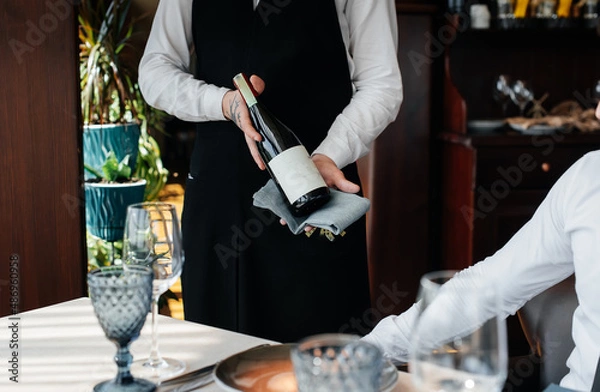 Fototapeta A young sommelier in a stylish uniform demonstrates and offers the client fine wine in the restaurant. Customer service. Table setting in a fine restaurant.