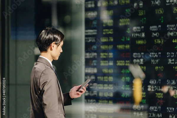 Obraz 株価ボードを見る男性（スマートフォンを手に持つ）