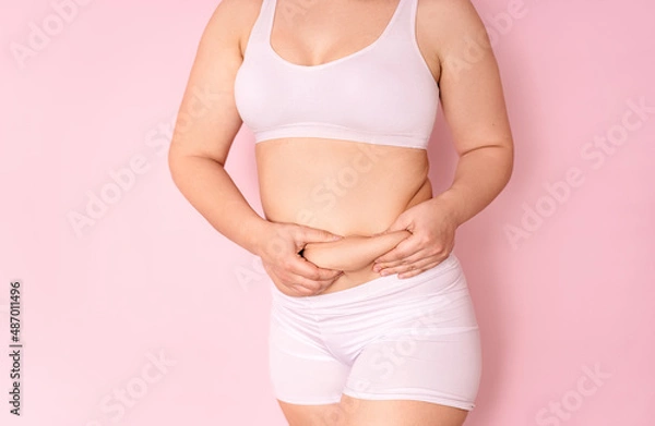 Fototapeta a fat girl holding a hand with excess fat on her stomach on a pink background. the concept of a woman's lifestyle with a diet to reduce the belly. recovery after childbirth. place for text
