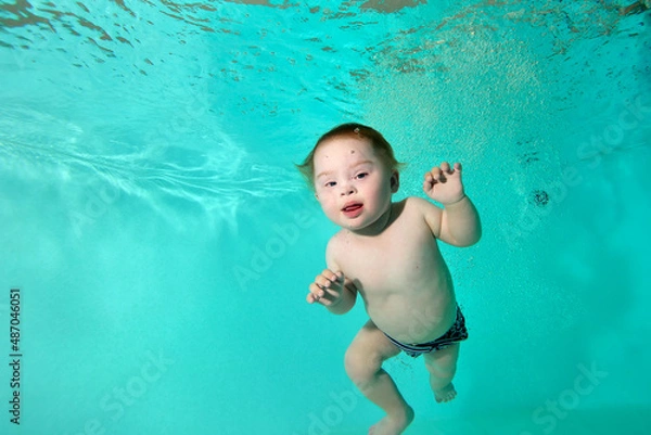 Obraz Portrait of a happy disabled child with Down syndrome who swims and plays underwater in a turquoise water pool. Close-up. Horizontal orientation.