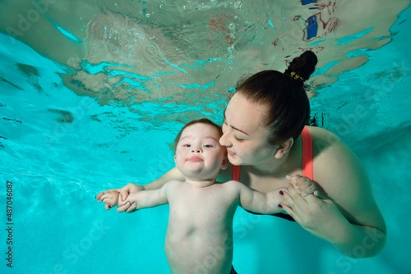 Obraz Happy Family: A mother and a child with Down syndrome enjoy swimming underwater in a turquoise water pool. The woman looks at the baby and smiles, holding his hands. Portrait. Horizontal orientation.
