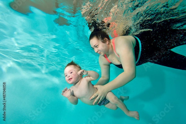 Obraz A happy mom and a child with Down syndrome swim together underwater in a turquoise water pool. The woman supports the baby with her hands and smiles. Portrait. Horizontal orientation.