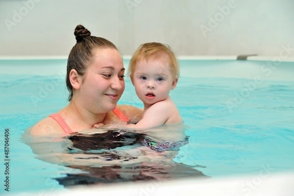 Obraz An adult woman holds a baby with Down syndrome in her arms while standing in the water in a children's pool with blue water. She looks at the child and smiles. Portrait. Horizontal orientation.