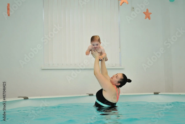 Obraz Mom raised and holds a baby with Down syndrome over her head, standing in the water in the pool. An infant swimming coach is engaged with a disabled child in a children's pool. Horizontal orientation.