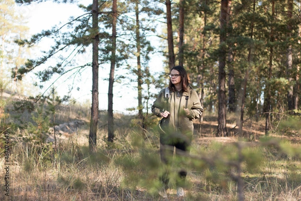 Fototapeta cute girl with dark long hair in glasses looks down standing in the forest with a backpack