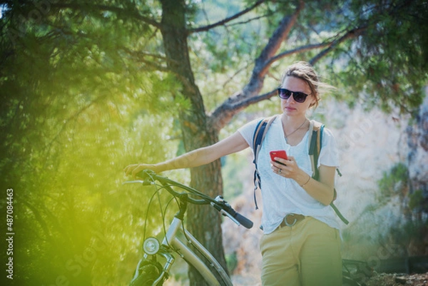 Obraz Young woman on a bicycle with a smartphone in her hands on a bike ride in the forest, summer travel and adventure