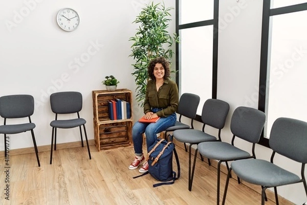 Fototapeta Young hispanic woman holding book sitting on chair at waiting room