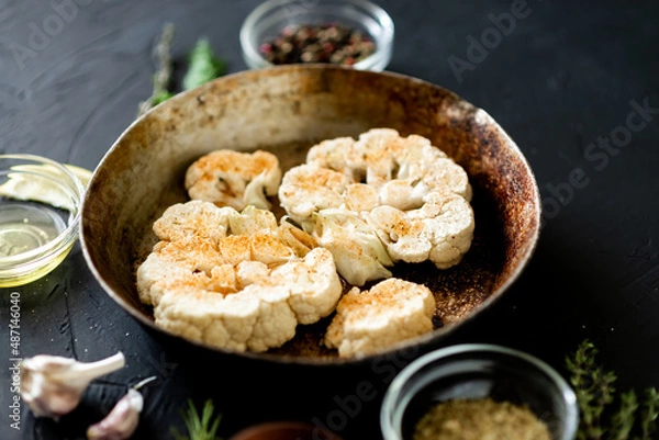 Obraz Cauliflower steak cooking. Raw cauliflower sprinkled with spices lies in a frying pan. Olive oil, herbs, various spices nearby. Dark background.