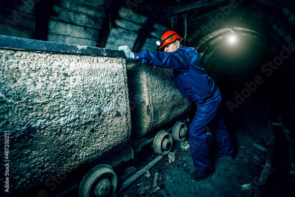 Fototapeta Tired miner in an old coal mine in the process of working. A miner in a blue protective suit with an orange helmet on his head. Copy space.