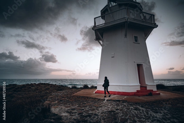Obraz Girl standing next to a lighthouse at sunset
