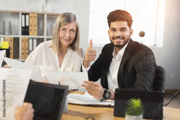 Fototapeta Young indian man and caucasian senior woman smiling and looking at camera during paperwork at office and showing thumb up. Two company workers in formal wear cooperating together.