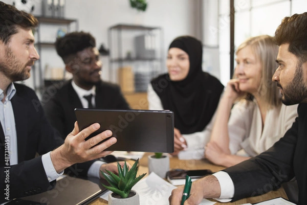 Fototapeta Multi ethnic team of business partners looking on one tablet screen during brainstorming at conference room. Five people discussing common working project at office.