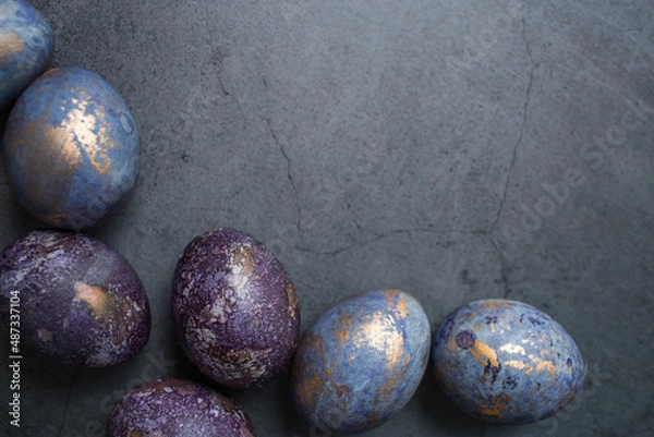 Fototapeta Easter card with a copy of the place for the text. Purple, blue and golden eggs on a dark background. The trend of 2022 purple shade is very peri. Natural dye karkade tea. Top view.