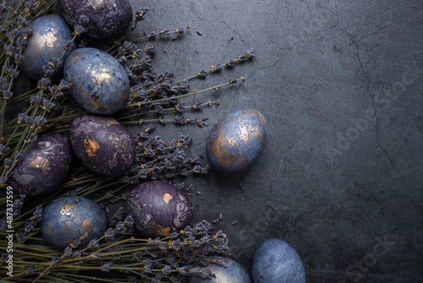 Fototapeta Easter card with a copy of the place for the text. Purple, blue and golden eggs with lavender on a dark background. The purple hue trend of 2022 is very relevant. Natural dye karkade tea. Top view.