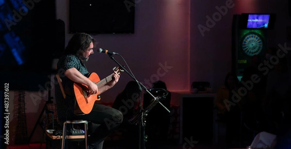 Fototapeta Singer-songwriter plays guitar and sings at acoustic concert in a club with an intimate atmosphere. Silhouettes of the public in the background