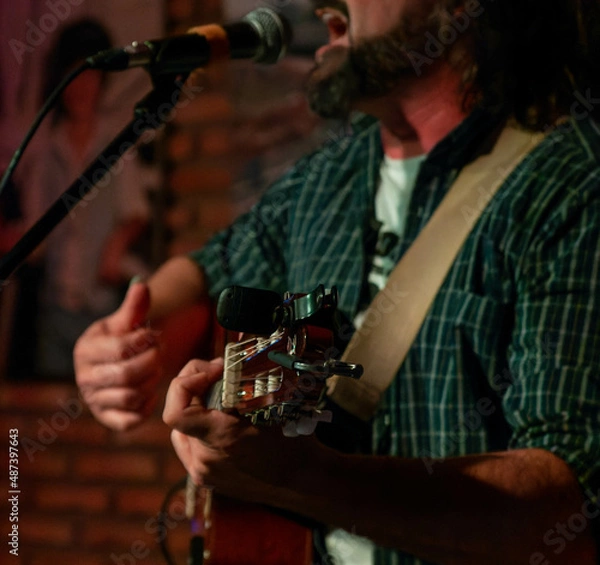 Fototapeta Singer-songwriter plays guitar and sings at acoustic concert in a club with an intimate atmosphere. . Exhibition of pictures  in the background